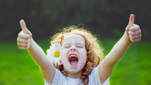 A little girl, with a daisy in her hair wearing a short sleeved light blue dress, with 2 thumbs up, on a sunny day with grass in the background.